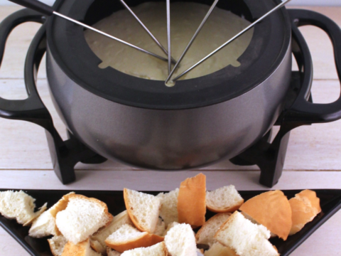 A fondue pot with several skewers is filled with melted cheese, perfect for delicious dips during football season. In front, a tray holds an assortment of cubed bread pieces for dipping. The setup rests invitingly on a light wooden surface.