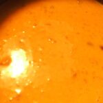 A close-up of a creamy orange soup or sauce, possibly with small bits of ingredients visible, resembles a decadent skillet queso recipe. The glossy surface, kissed by light reflections, exudes a rich and inviting appearance.