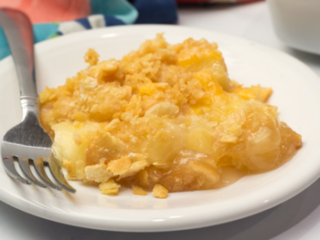 A slice of pineapple crumble dessert on a white plate with a fork showcases one of the delightful fresh pineapple recipes. A colorful cloth in the background sits beside a white baking dish filled with more of this tropical treat.