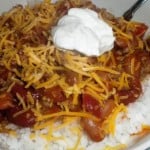 A bowl of saucy beef chili served over white rice, topped with shredded cheddar cheese and a dollop of sour cream. The hearty chili contains kidney beans, ground meat, and diced tomatoes. A spoon is partially visible in the bowl, ready to dive into this delicious recipe.