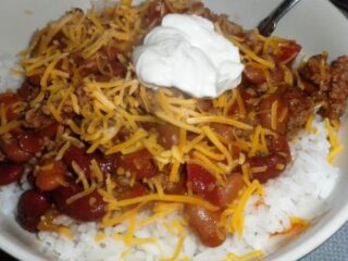 A bowl of saucy beef chili served over white rice, topped with shredded cheddar cheese and a dollop of sour cream. The hearty chili contains kidney beans, ground meat, and diced tomatoes. A spoon is partially visible in the bowl, ready to dive into this delicious recipe.