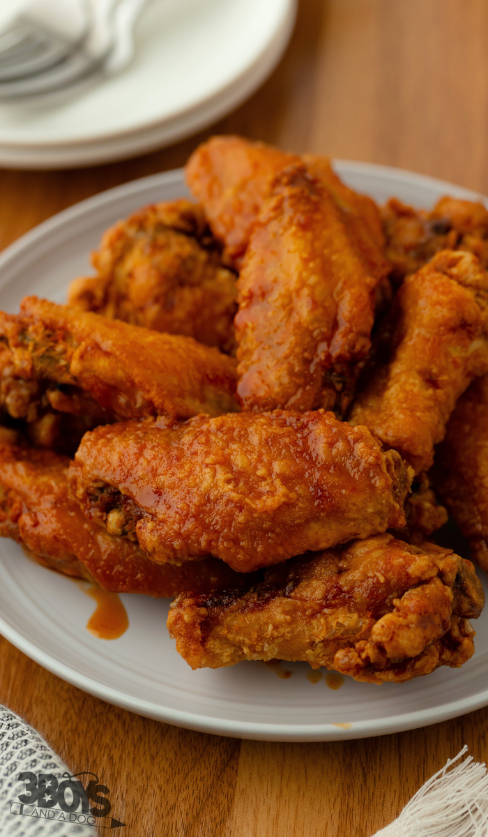 A plate of crispy, golden-brown buffalo chicken wings piled high on a white dish, reminiscent of an East Asian Style Wings recipe, set on a wooden table. In the background, part of another small white plate with utensils is visible, slightly out of focus.