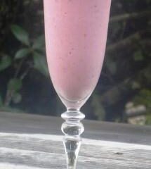 A tall, elegant glass filled with a creamy strawberry smoothie sits on a wooden surface. The smoothie, following a delightful recipe, is garnished with a small, heart-shaped piece of fruit on the rim. In the background, blurred greenery adds a serene touch.