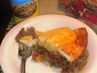 A plate with a slice of Impossible Taco Pie featuring ground meat and a cheesy crust. A fork rests on the plate, and nearby sits a container of taco seasoning alongside a fringed cloth.