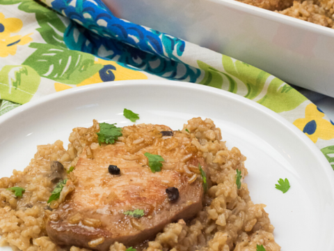 A plate of baked pork chops with seasoned rice, beautifully garnished with herbs, rests on a colorful tablecloth. In the background, a casserole dish showcases the tempting pork chop and rice casserole recipe for more deliciousness.