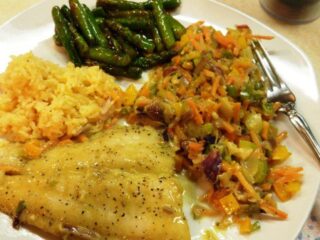 A plate of food featuring a Thai-inspired grilled catfish fillet, a side of sautéed mixed vegetables, rice, and asparagus. A fork and knife are placed on the side of the plate, adding to this delightful coconut-infused feast.