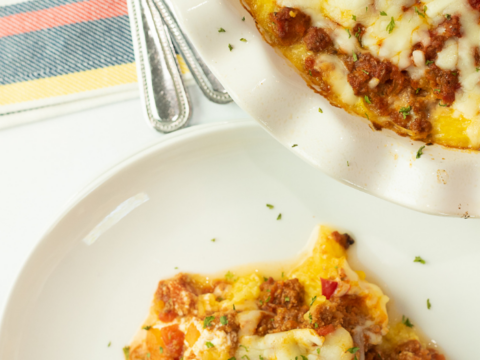 Top view of a dish featuring melted cheese and meat sauce over delectable spaghetti squash. A serving sits on a white plate beneath it, while forks rest on a colorful striped cloth nearby. A logo is tucked in the bottom left corner, completing this delicious presentation.