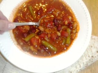 A bowl of hot soup containing red kidney beans, green beans, tomatoes, and other vegetables sits invitingly. A hand holding a spoon is ready to scoop up this delicious recipe, while the wooden table and lace cloth complete the cozy scene.