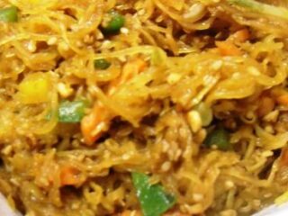 A close-up of a dish featuring shredded vegetables, including green and orange pieces, mixed with thin noodles. Enhanced with a nutty flavor, the dish showcases spaghetti squash and visible sesame seeds, giving it a textured and appetizing look.