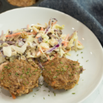 A white plate with two savory meat muffins garnished with herbs is perfect for brunch, served alongside a creamy coleslaw with shredded carrots and cabbage. In the background, a muffin tray hints at more delicious mini muffin recipes waiting to be discovered on the dark cloth napkin.