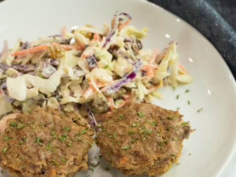 A white plate with two savory meat muffins garnished with herbs is perfect for brunch, served alongside a creamy coleslaw with shredded carrots and cabbage. In the background, a muffin tray hints at more delicious mini muffin recipes waiting to be discovered on the dark cloth napkin.