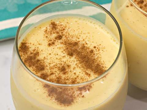 Two glasses of creamy yellow eggnog, reminiscent of easy banana dessert recipes, topped with a sprinkle of cinnamon on a white surface. A blue napkin with floral patterns is partially visible in the background.
