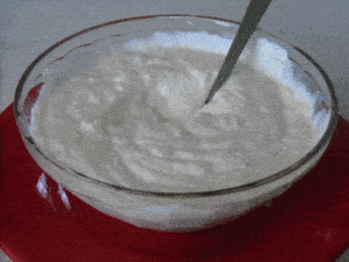 A spoon is rapidly stirring thick, creamy Parmesan dip in a transparent bowl placed on a red surface. The dips texture is smooth, and the motion causes it to swirl around vigorously.