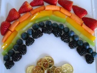 A delightful Rainbow Fruit Plate Recipe features a vibrant medley of strawberries, cantaloupe, pineapple, green apple slices, blackberries, and blueberries on a white background, crowned with gold foil-wrapped chocolate coins at the base.