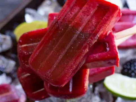 A stack of red popsicles on wooden sticks is arranged in a glass surrounded by ice on a dark surface. Fresh blackberries and lime slices, reminiscent of refreshing summer recipes, are scattered around, adding vibrant colors to the scene.