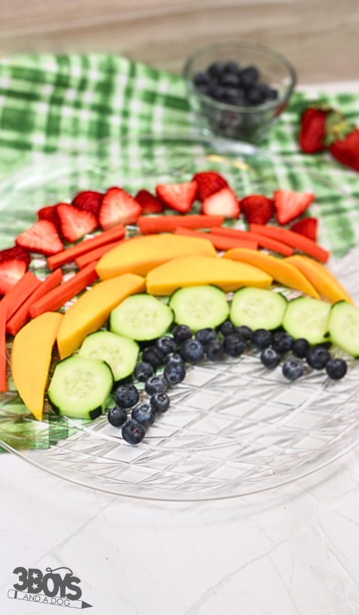 A clear plate showcases a vibrant Rainbow Fruit and Vegetable Plate Recipe. From top to bottom: strawberries, carrots, mango slices, cucumbers, and blueberries. The plate rests on a green and white checked cloth with whole strawberries and blueberries nearby.