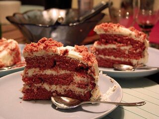 Slices of rich red velvet cake with creamy white frosting are served on plates, ready to savor with spoons. In the blurred background on a wooden table, glasses and a bowl set the scene for this delightful recipe moment.