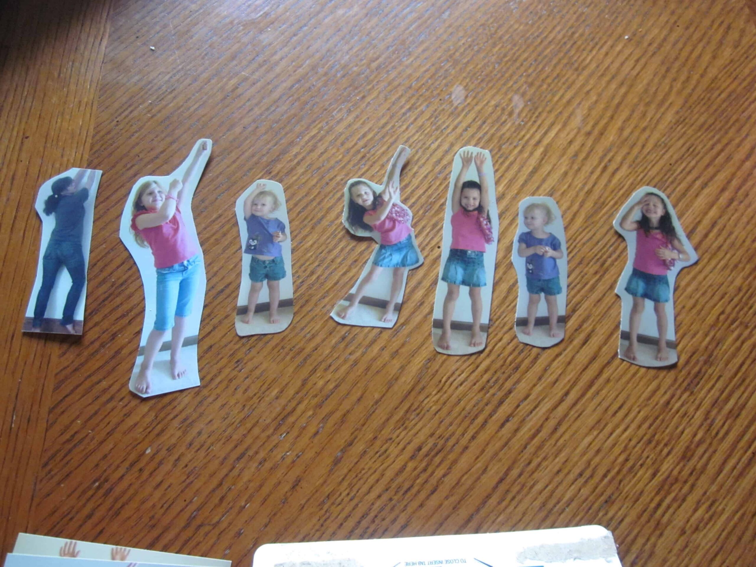 Cutout images of a child in various playful poses are arranged on a wooden surface, wearing a pink top and blue skirt. Perfect for back-to-school bookmarks, these lively depictions capture the essence of familiar faces in every stance. A cutout of the child turned away is seen on the left.
