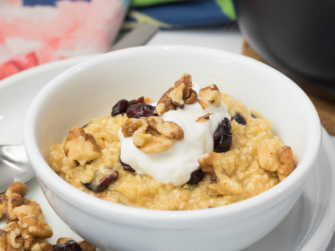 A delicious breakfast recipe awaits: a bowl of oatmeal topped with walnuts, dried cranberries, and a dollop of yogurt sits on a white plate. Additional walnuts and cranberries surround it. A colorful cloth adds charm to the scene, with a pot partially visible in the background.