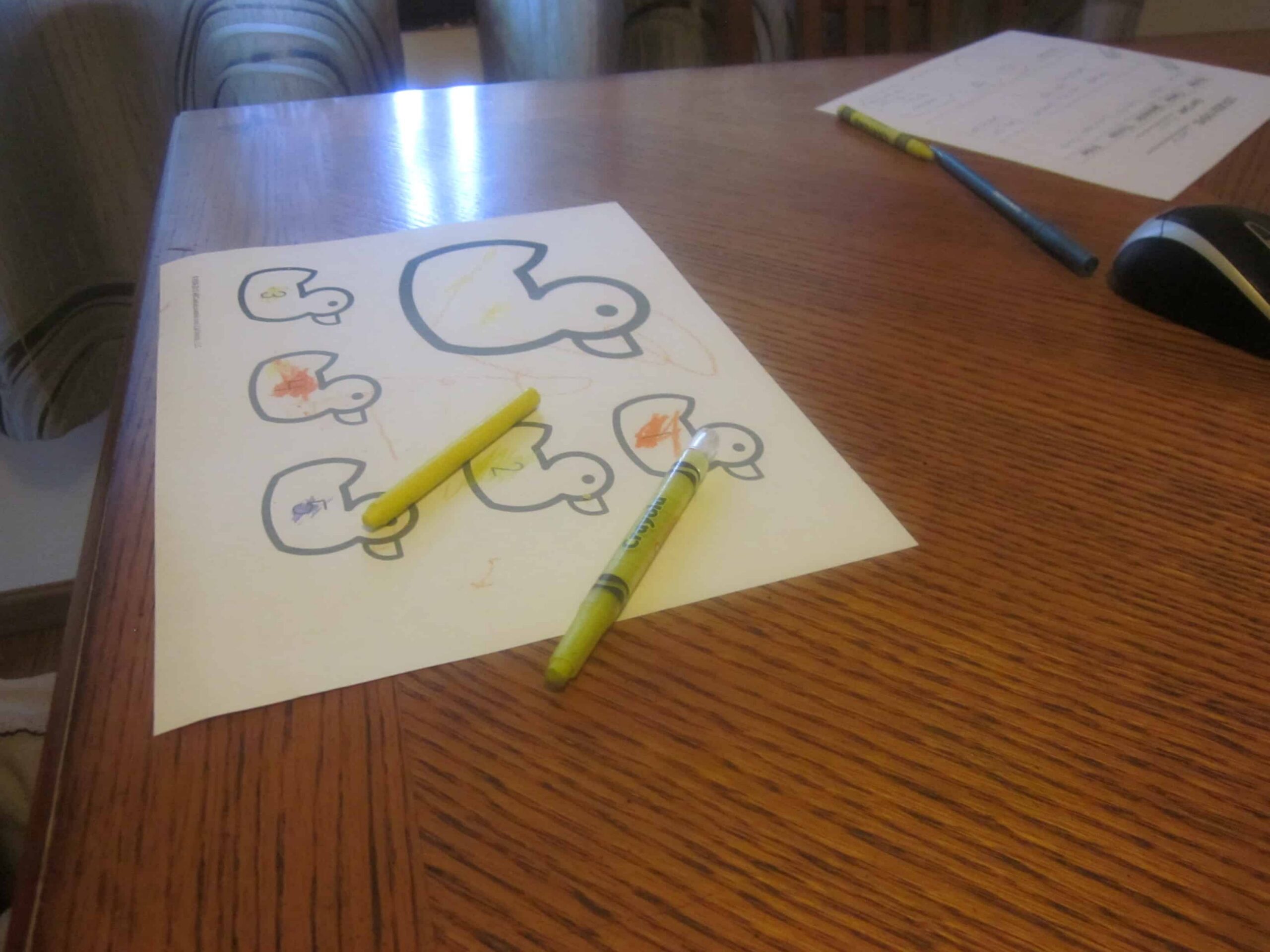 A wooden table hosts a coloring sheet with outlines of ducks, perfect for a preschool activity. Two yellow crayons, one capped and one uncapped, assist in bringing the Five Little Ducks to life. In the background lie a black pen and paper, ready for counting practice.