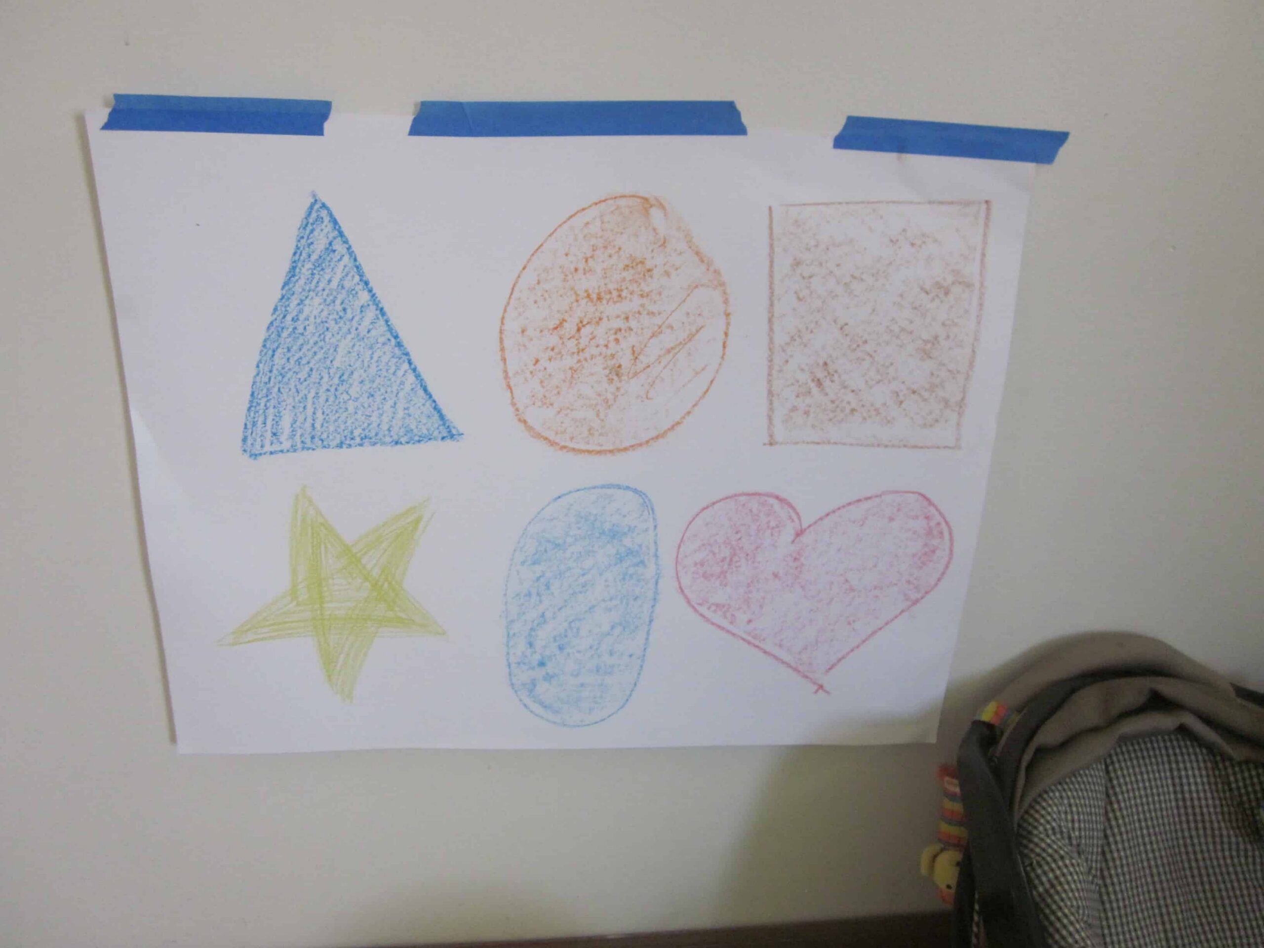 Hand-drawn shapes on paper taped to a wall include a blue triangle, orange circle, orange square, yellow star, blue oval, and red heart. A portion of a chair is visible at the bottom right—turning simple shapes into fun and engaging activities.