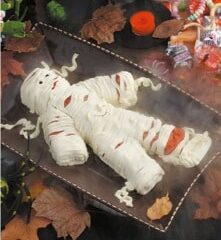 A yummy mummy pastry lies on a brown tray, surrounded by autumn leaves and Halloween decorations like a skeleton and candies. Wrapped in white strips resembling bandages, this delightful creation is more than just decoration—its a spook-tacular Halloween recipe to savor.