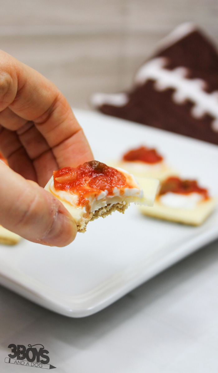 A hand holds a square cracker topped with cheese, tomato sauce, and herbs, a bite missing from this Southwestern Triscuit Bites recipe. In the background, a white plate displays more of these tasty snacks beside a folded brown and white patterned cloth.