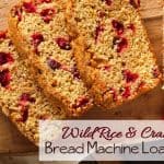Slices of wild rice and cranberry bread loaf rest on a wooden board, showcasing a golden crust with cranberries throughout. Text overlay reads Wild Rice & Cranberry Bread Machine Loaf Recipe.
