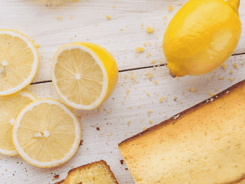 Sliced lemon cake on a white wooden surface, with fresh lemon slices and whole lemons nearby. A yellow fabric is in the background. The text reads Try our Bread Machine Lemon Cake recipe today!.