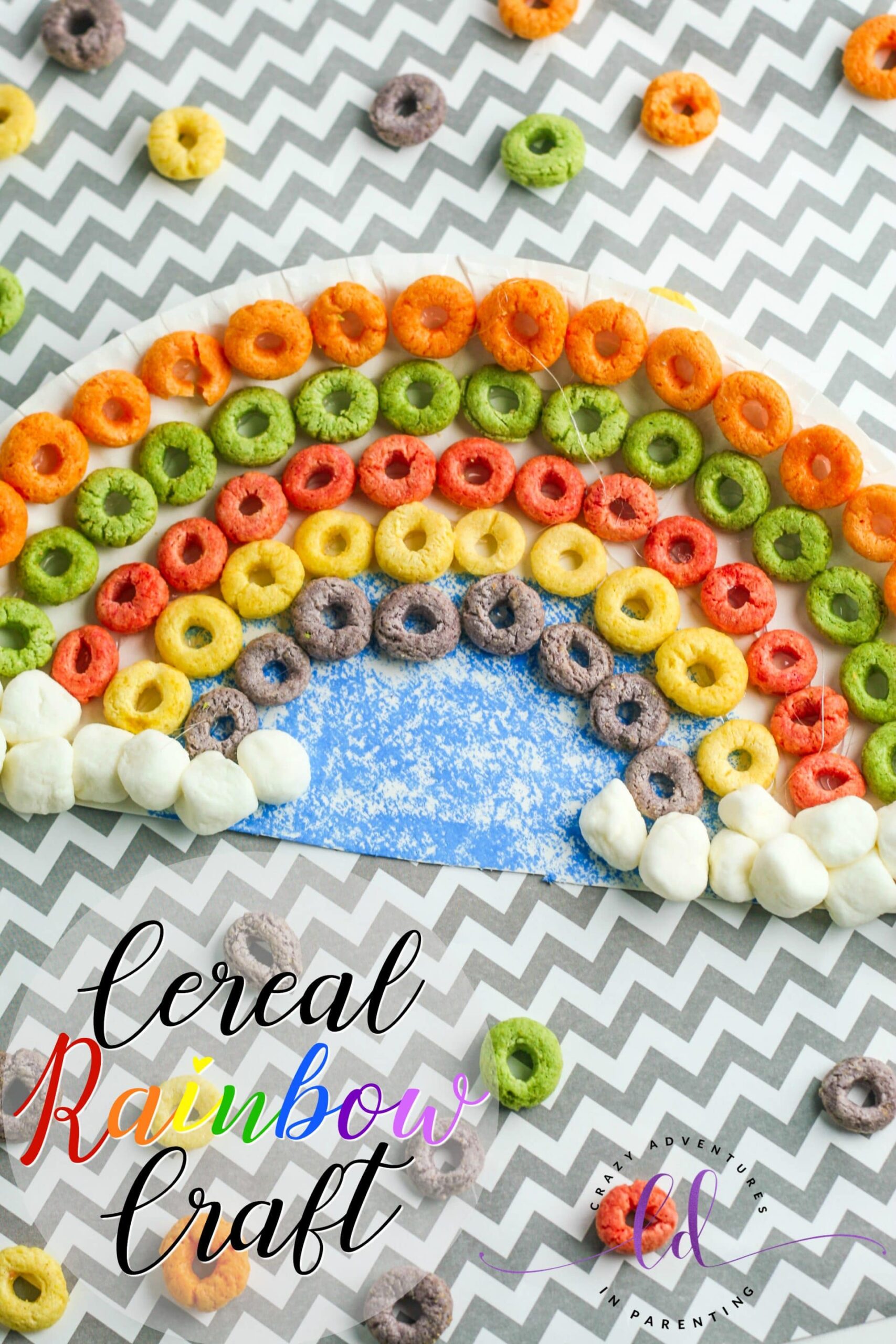 Colorful cereal rings are arranged in a rainbow pattern on zigzag paper, crafting a vibrant metaphor for finding joy at the end of the rainbow. Cotton balls at each end enhance this delightful scene. The text reads Cereal Rainbow Craft in a playful font.