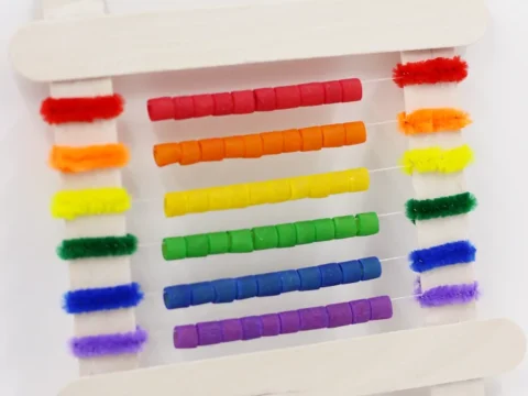 A homemade rainbow abacus, crafted with colorful beads and pipe cleaners, stretches across two large craft sticks, showcasing vibrant hues from red to purple. Above the abacus, the phrase End of the Rainbow is playfully written in a burst of colors.
