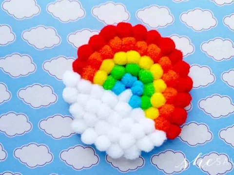 A colorful rainbow made of small, fluffy pom-poms sits on a blue background with white cloud patterns, as if you could find it at the end of the rainbow.