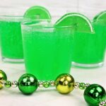 Three clear plastic cups filled with fizzy green drinks, each garnished with a lime wedge, hint at a festive St. Patricks Day mocktail recipe. In the foreground, a green and gold beaded necklace adds to the celebratory spirit on a crisp white surface.