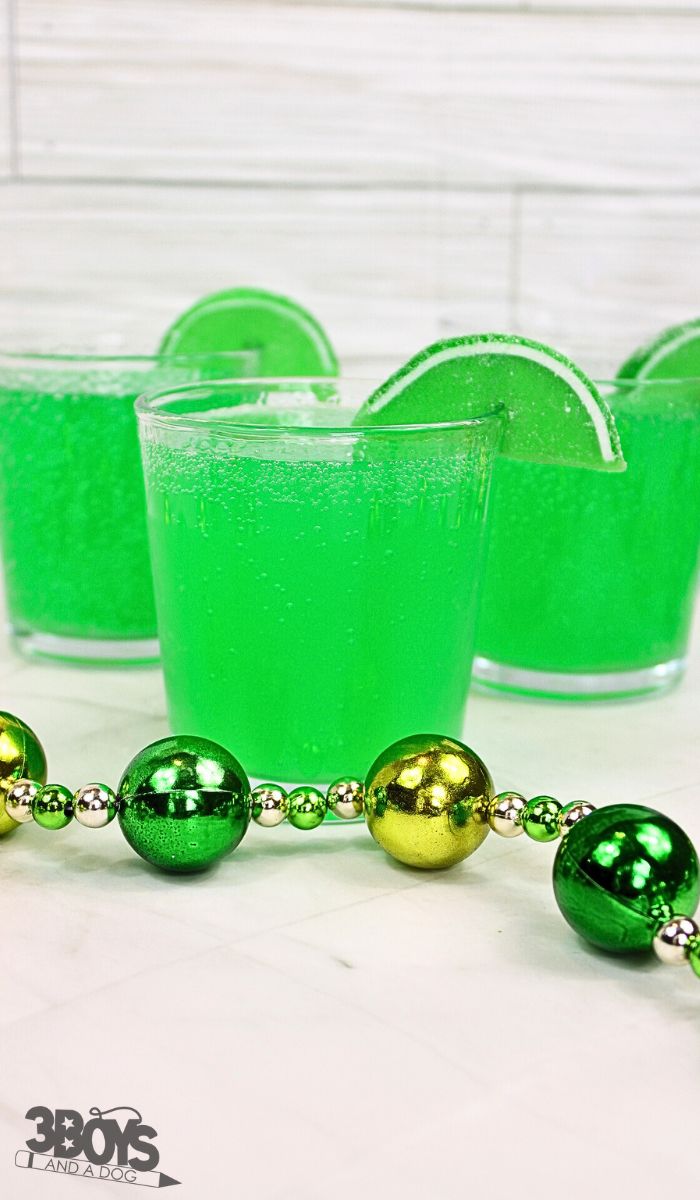 Three clear plastic cups filled with fizzy green drinks, each garnished with a lime wedge, hint at a festive St. Patricks Day mocktail recipe. In the foreground, a green and gold beaded necklace adds to the celebratory spirit on a crisp white surface.