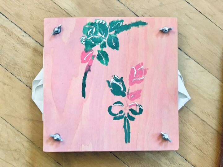A square wooden piece adorned with sweet pink roses and green leaves is elegantly painted on the surface. Four metal bolts anchor each corner. Resting on a wood-grained floor, this charming piece makes a perfect Valentine favor.