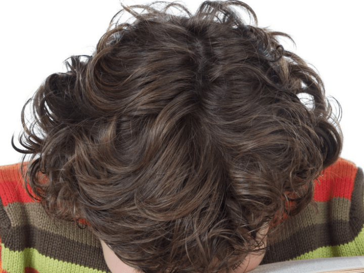 A boy with curly hair rests his head on an open book, embracing the learning curve. The text above reads, "5 Unexpected Ways Boys Learn." He wears a colorful striped sweater.