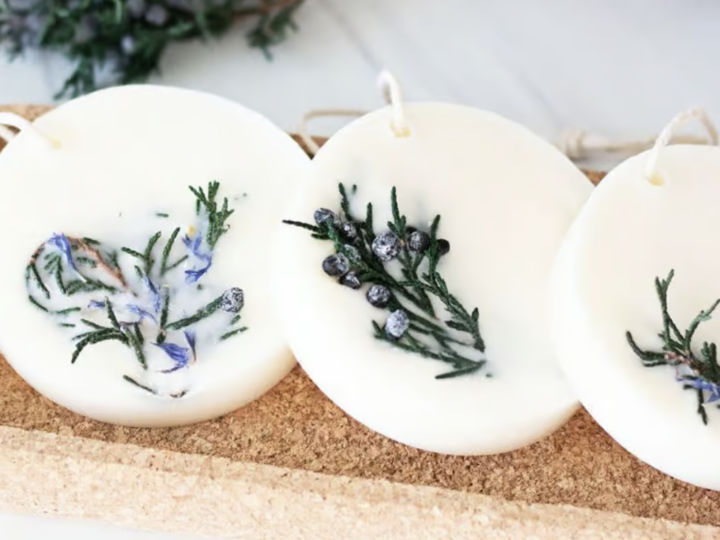 Three round white wax tablets with sprigs of green rosemary and small berries rest on a rectangular cork tray, embodying the spirit of National Craft Month. Each tablet features a string loop for hanging, with soft lighting highlighting their natural charm against a blurred background.