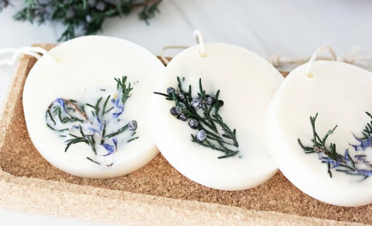 Three round white wax tablets with sprigs of green rosemary and small berries rest on a rectangular cork tray, embodying the spirit of National Craft Month. Each tablet features a string loop for hanging, with soft lighting highlighting their natural charm against a blurred background.