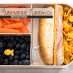 A bento box featuring a submarine sandwich with ham and cheese, baby carrots, juicy blueberries crowned with an orange fish cracker, and a side of orange fish-shaped crackers. Perfect for those seeking bento-style lunches for adults. Text reads Submarine Sandwich Bento Box Recipe.