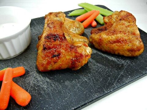 Grilled Chipotle Chicken Wings rest on a black slate board, accompanied by baby carrots and celery sticks. A small white ramekin, likely with dipping sauce, sits beside the wings.