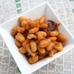 A square white bowl brimming with glossy baked beans in a rich sauce sits on a tiled surface. The beans, perfectly cooked, are complemented by small bits of bacon that add a savory touch to the dish.