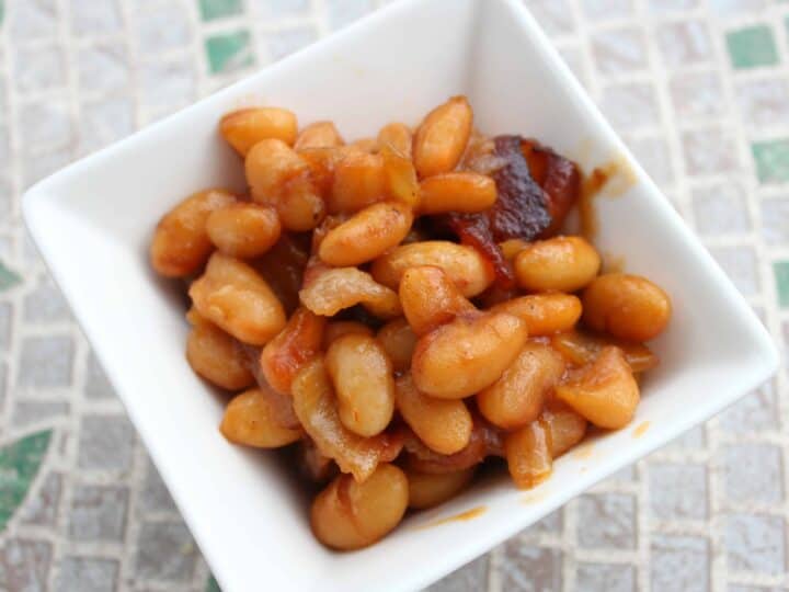 A square white bowl brimming with glossy baked beans in a rich sauce sits on a tiled surface. The beans, perfectly cooked, are complemented by small bits of bacon that add a savory touch to the dish.