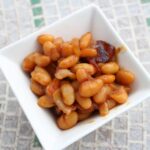 A square white bowl brimming with glossy baked beans in a rich sauce sits on a tiled surface. The beans, perfectly cooked, are complemented by small bits of bacon that add a savory touch to the dish.