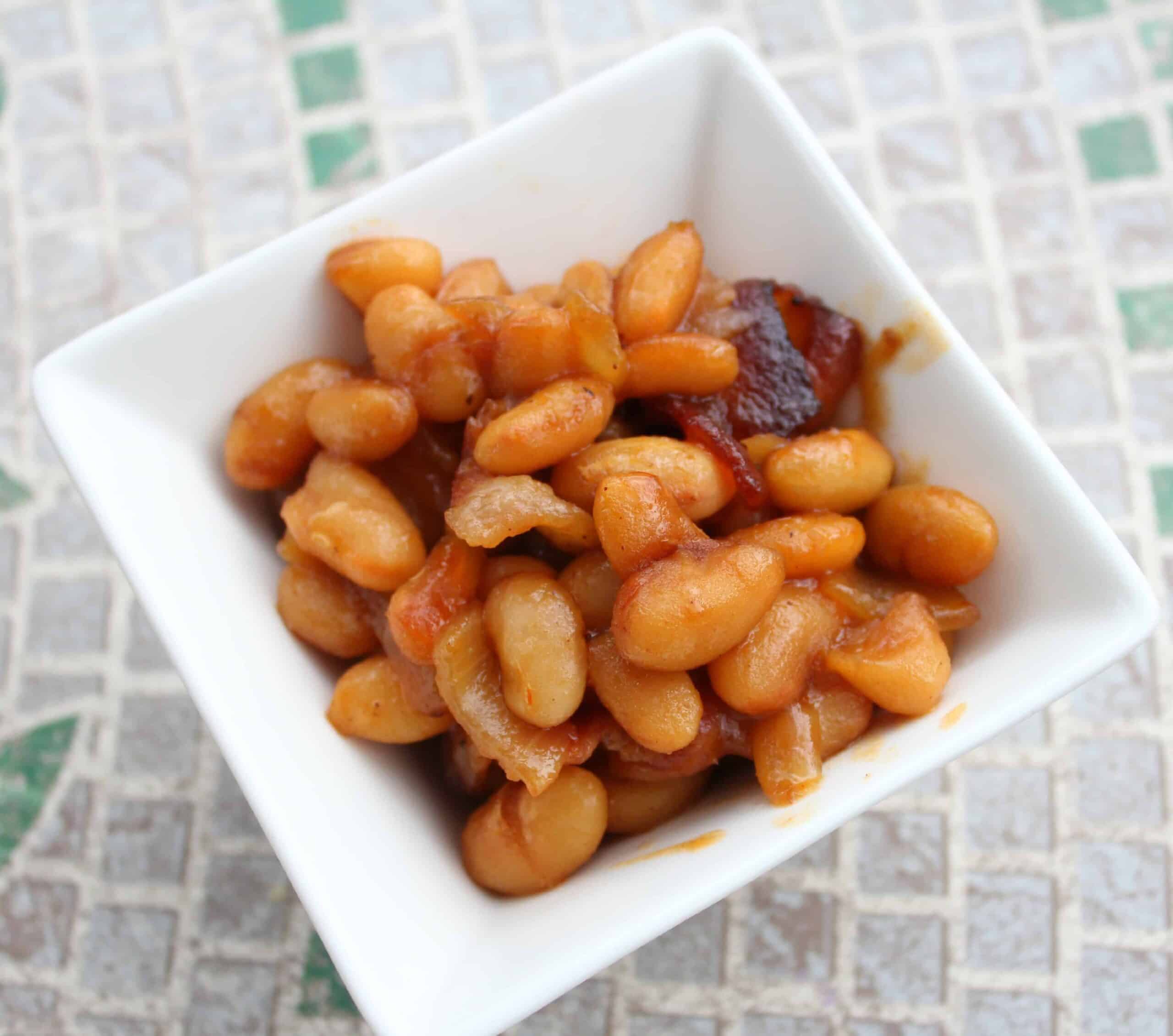 A square white bowl brimming with glossy baked beans in a rich sauce sits on a tiled surface. The beans, perfectly cooked, are complemented by small bits of bacon that add a savory touch to the dish.
