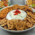 A platter of round pretzel crisps surrounds a bowl of creamy Garlic Cheese Dip topped with red pepper pieces. In the background, two bags of pretzel crisps are visible, showing images of the snack.