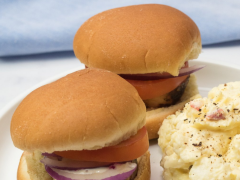 Two savory hamburgers with tomato and red onion slices are placed on a white plate next to a serving of potato salad, reminiscent of favorite pork recipes. A blue and white cloth is in the background, adding a touch of elegance.