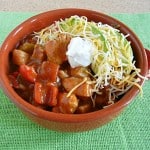 A bowl of Chicken Fajita Chili brimming with diced vegetables and chunks of meat, topped with shredded cheese and a dollop of sour cream, sits invitingly on a green placemat.