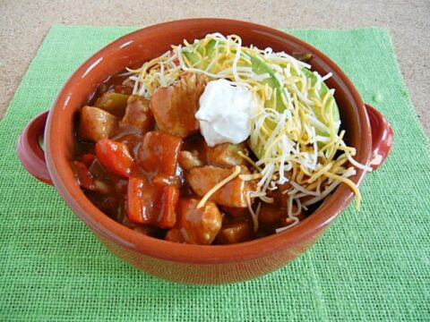 A bowl of Chicken Fajita Chili brimming with diced vegetables and chunks of meat, topped with shredded cheese and a dollop of sour cream, sits invitingly on a green placemat.