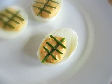Close-up of tailgating deviled eggs on a white plate. Each egg half is topped with crisscrossed green chives over the yellow filling, perfect for any gathering. Three egg halves are visible.