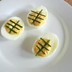 Three tailgating deviled eggs are arranged on a white plate. Each egg is halved with a creamy filling and garnished with two crossed chives on top, creating a decorative pattern. The plate is set on a cork surface, ready to impress your game-day guests.